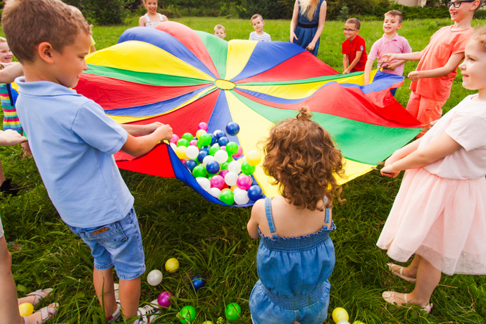 Gruppe von Kindern spielt im Freien auf einer Wiese mit einem bunten Schwungtuch, in dem viele kleine Plastikbälle liegen, die durch die Bewegung in die Luft geworfen werden.
