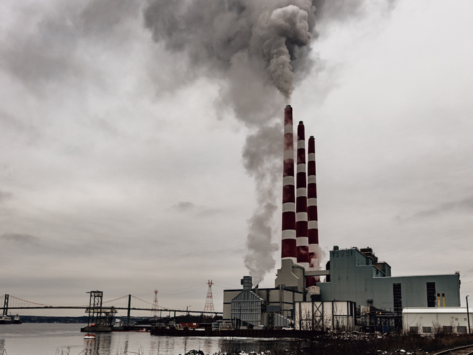 Industrieanlage mit mehreren hohen Schornsteinen, aus denen dichter Rauch in den grauen Himmel aufsteigt, am Ufer eines Gewässers.