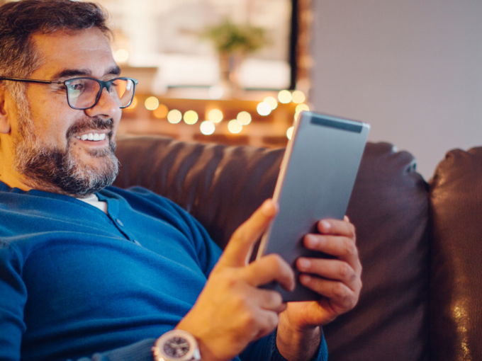 Eine lächelnde Person sitzt mit einem Tablet auf dem Sofa.