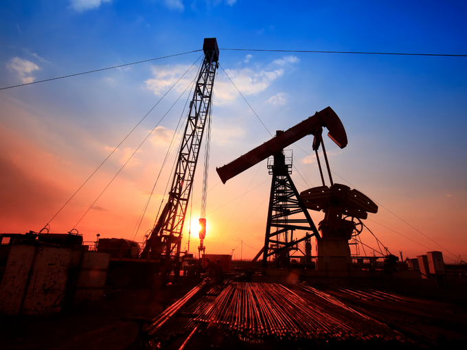 Silhouette einer Ölförderanlage mit Pumpjack und Bohrturm auf einem Industriegelände bei Sonnenuntergang, mit Rohrleitungen im Vordergrund und farbigem Himmel im Hintergrund.