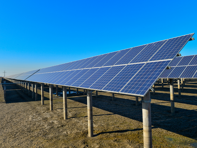 Großflächiger Solarpark mit mehreren Reihen schräg montierter Photovoltaikmodule auf Stützpfosten in einer trockenen, offenen Landschaft unter klarem blauem Himmel.