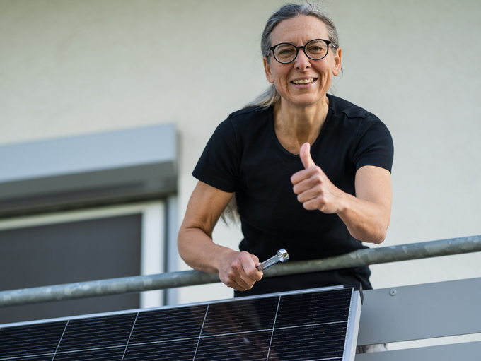 Person auf einem Balkon mit Solarmodul, die mit einem Werkzeug arbeitet und den Daumen hebt.