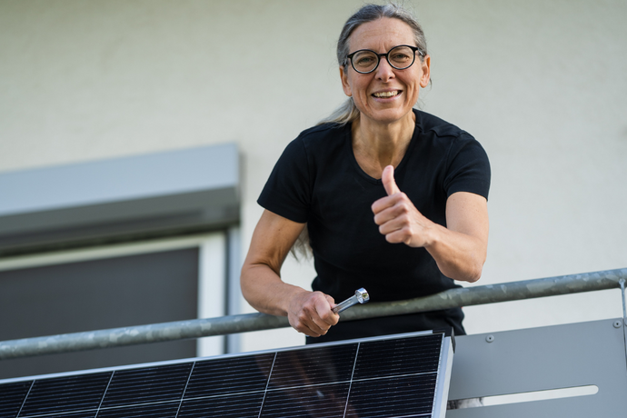 Person auf einem Balkon mit Solarmodul, die mit einem Werkzeug arbeitet und den Daumen hebt.